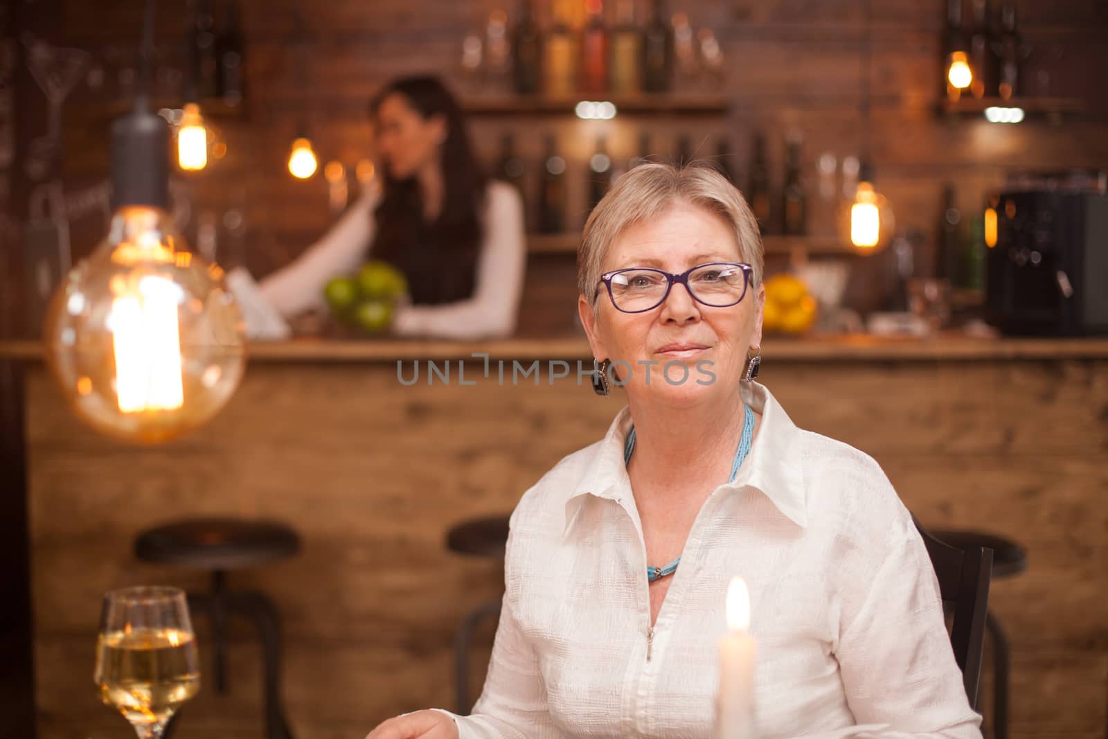 Portrait of elderly woman in a vintage restaurant. A glass of white wine. Blurred barmaid working in the background.