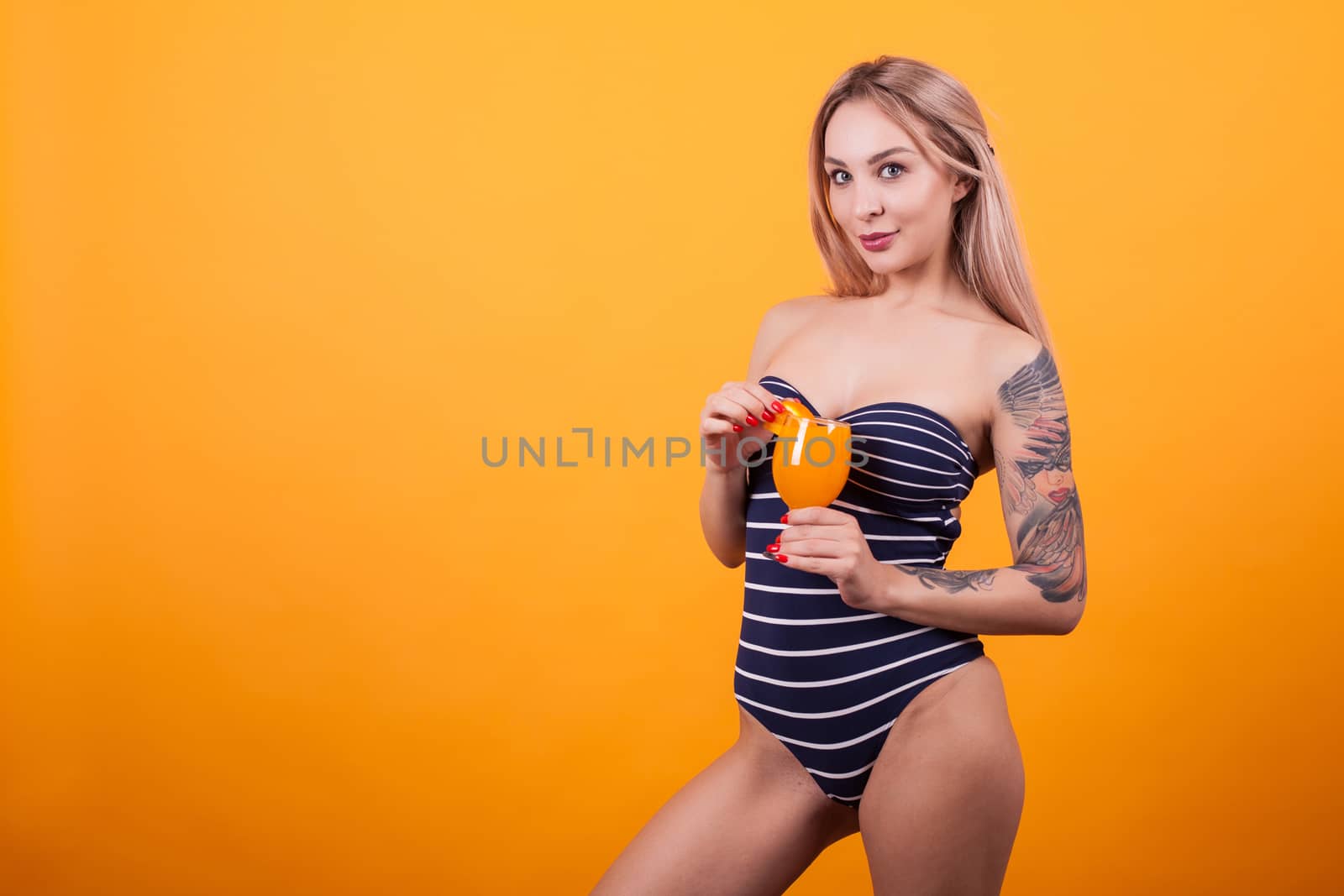 Beautiful young woman with tatto on her armn holding tasty drink over yellow background. Sexy woman. Delicious drink. Summer time.