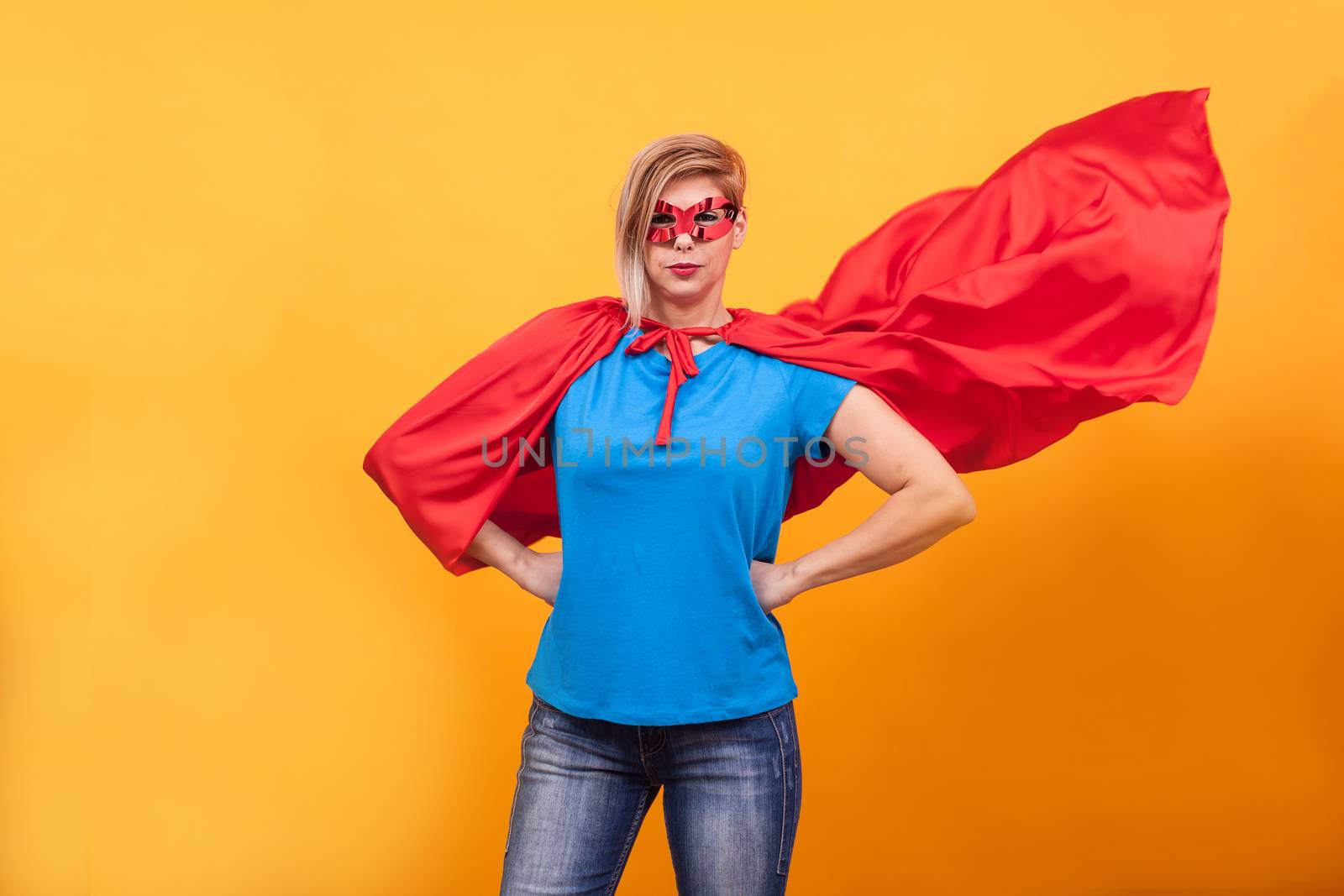 Young woman in superheros costume standing proudly with her red cape in the air over yellow background. Super powers.
