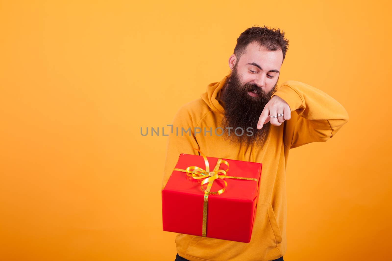Handsome bearded man in yellow hoodie pointing on red gift box over yellow background. Handsome man. Surprise gift.