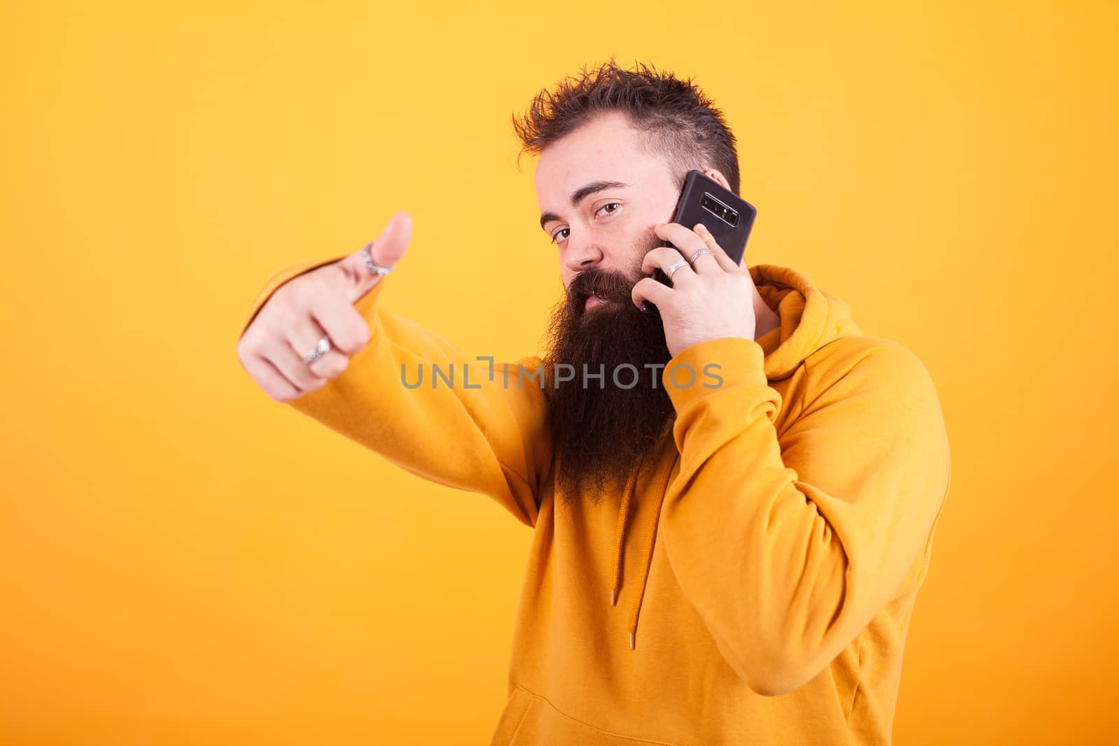 Attractive young man with long beard talking on the phone and showing thumbs up sign over yellow background. Hipster. Hand sign. Hoodie