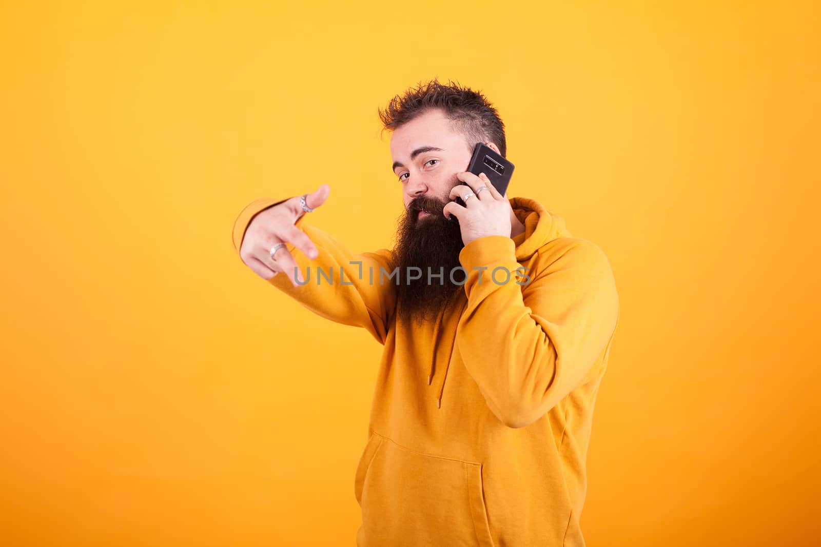 Trendy man wearing yellow hoddie talking on the phone and showing ok sign over yellow background. Smartphone. Stylish man.