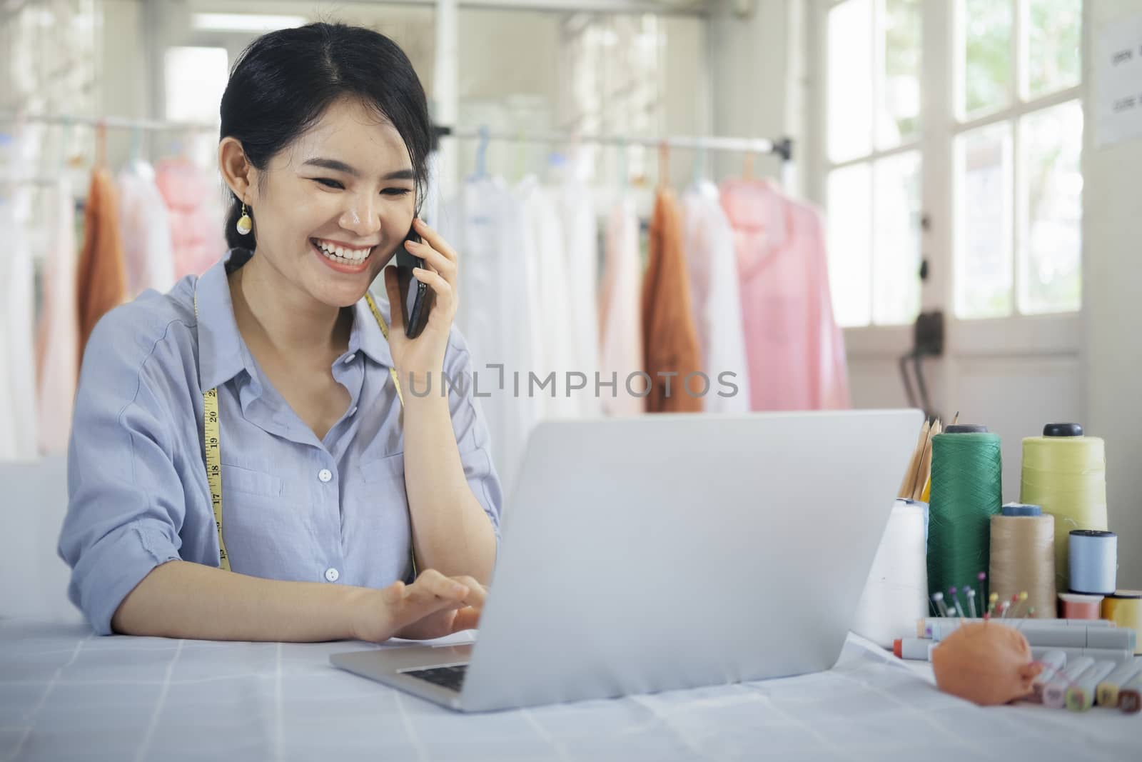 Young tailor design owner woman talking with customer on smart phone in her studio with clothes hanging in the background. Business owner shop and entrepreneur concept.