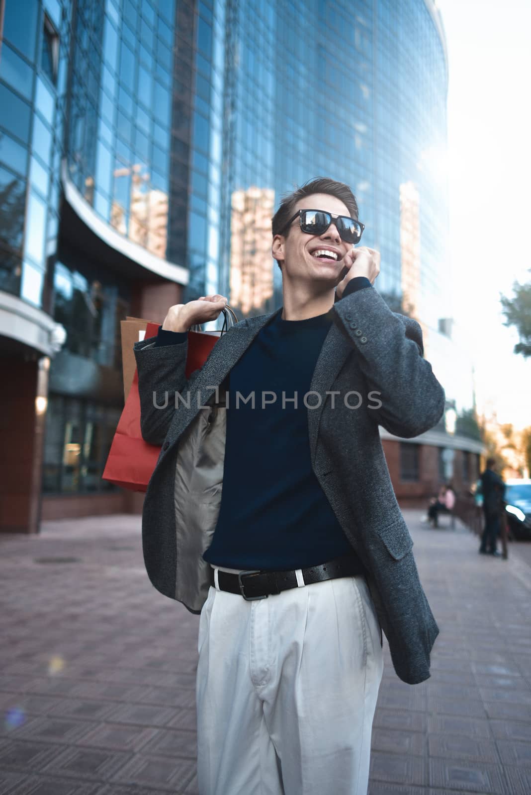 Happy stylish young man with phone walking in urban street and enjoying Black Friday shopping in trendy stores in city. by Nickstock