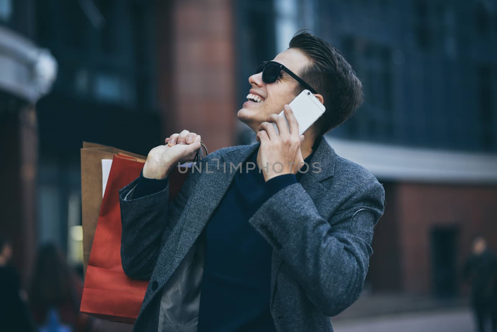 Happy stylish young man with phone walking in urban street and enjoying Black Friday shopping in trendy stores in city. by Nickstock