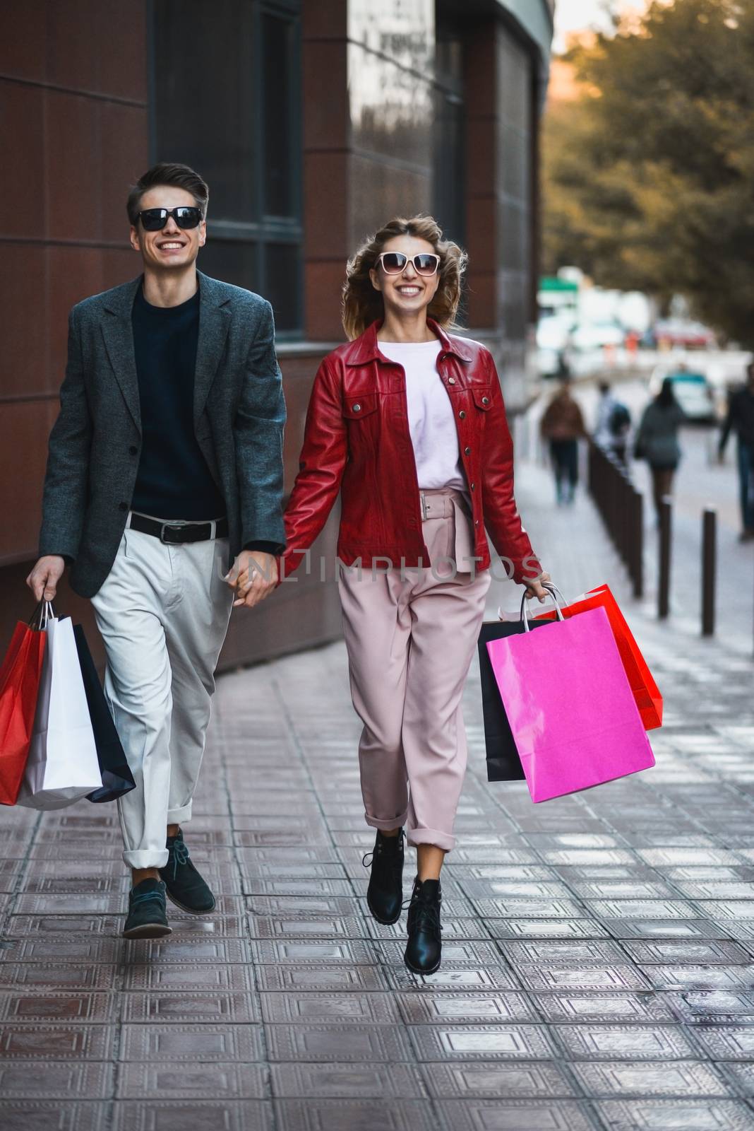 Front view of a casual couple of shoppers running in the street towards camera holding colorful shopping bags. by Nickstock