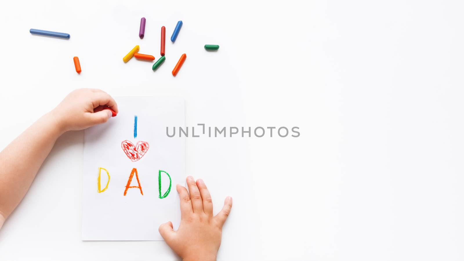 Left-handed toddler draws greeting card for Father's day. Kid uses wax crayons to write I Love Dad and to paint red heart symbol.Top view on child's hands,pencils on white background with copy space.