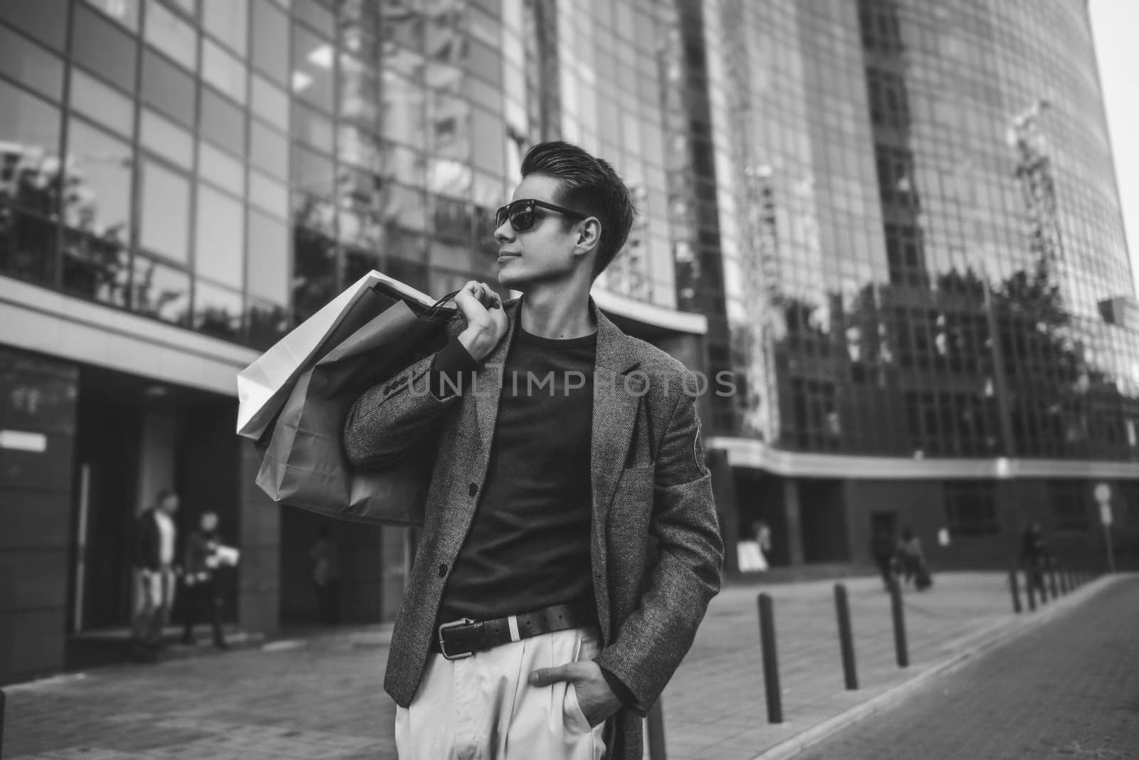 Serious stylish young man with sunglasses walking in urban street and enjoying Black Friday shopping in trendy stores in city by Nickstock