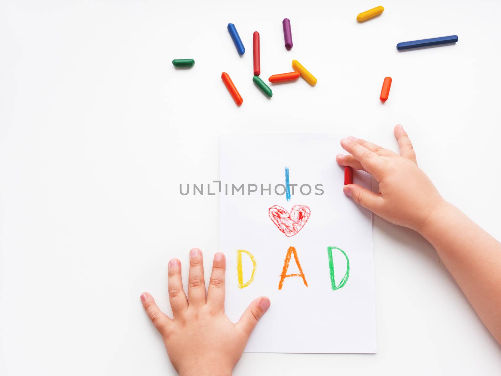 Toddler draws greeting card for Father's day. Kid uses wax crayons to write I Love Dad and to paint red heart symbol. Top view on child's hands and pencils on white background with copy space.
