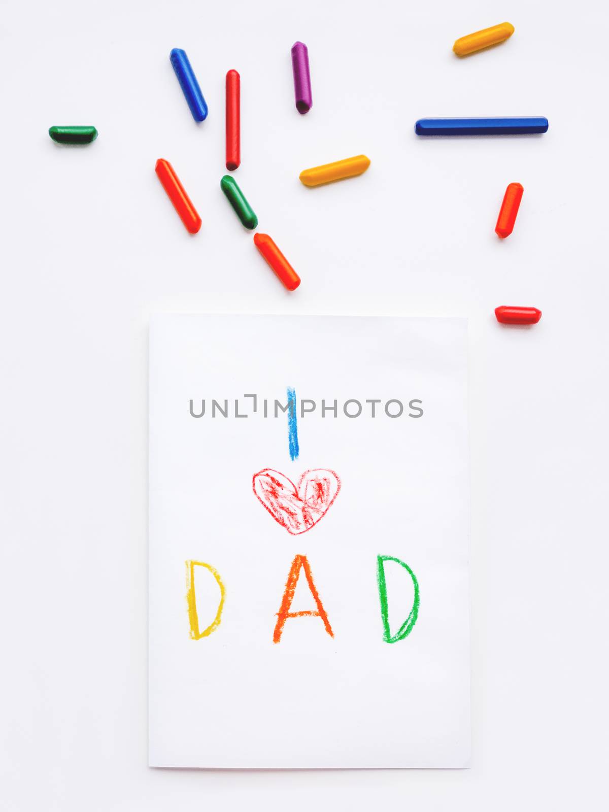 Top view on hand drawn greeting card for Father's day. Kid uses wax crayons to write I Love Dad and to paint red heart symbol. Flat lay with pencils on white background.