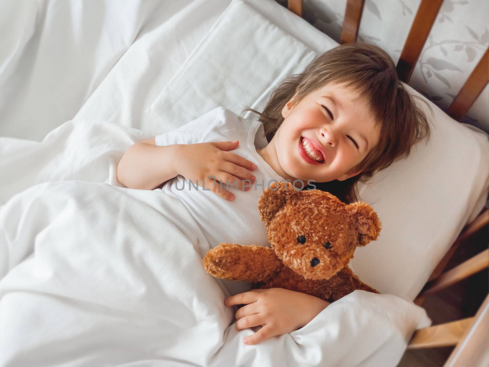 Toddler lies in bed with cute teddy bear. Little boy under white blanket with fluffy toy. Plush guard watches out child's sleep. Morning bedtime at cozy home.
