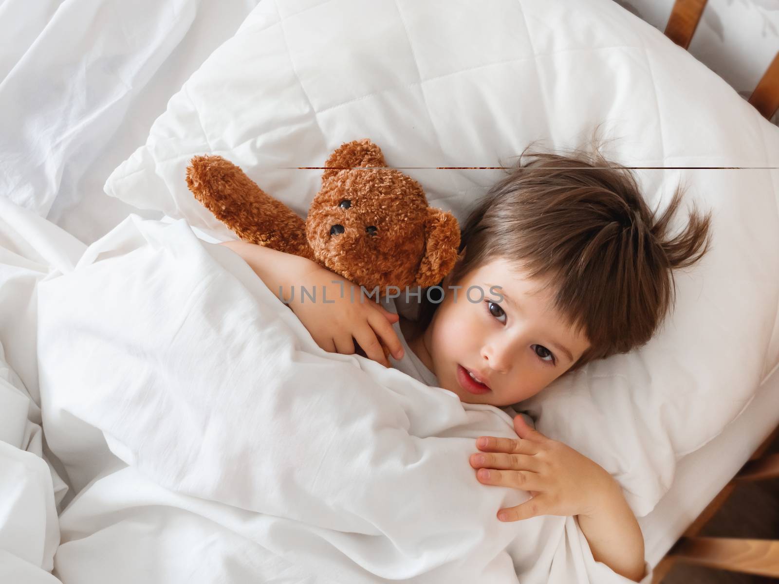 Toddler lies in bed with cute teddy bear. Little boy under white blanket with fluffy toy. Plush guard watches out child's sleep. Morning bedtime at cozy home.