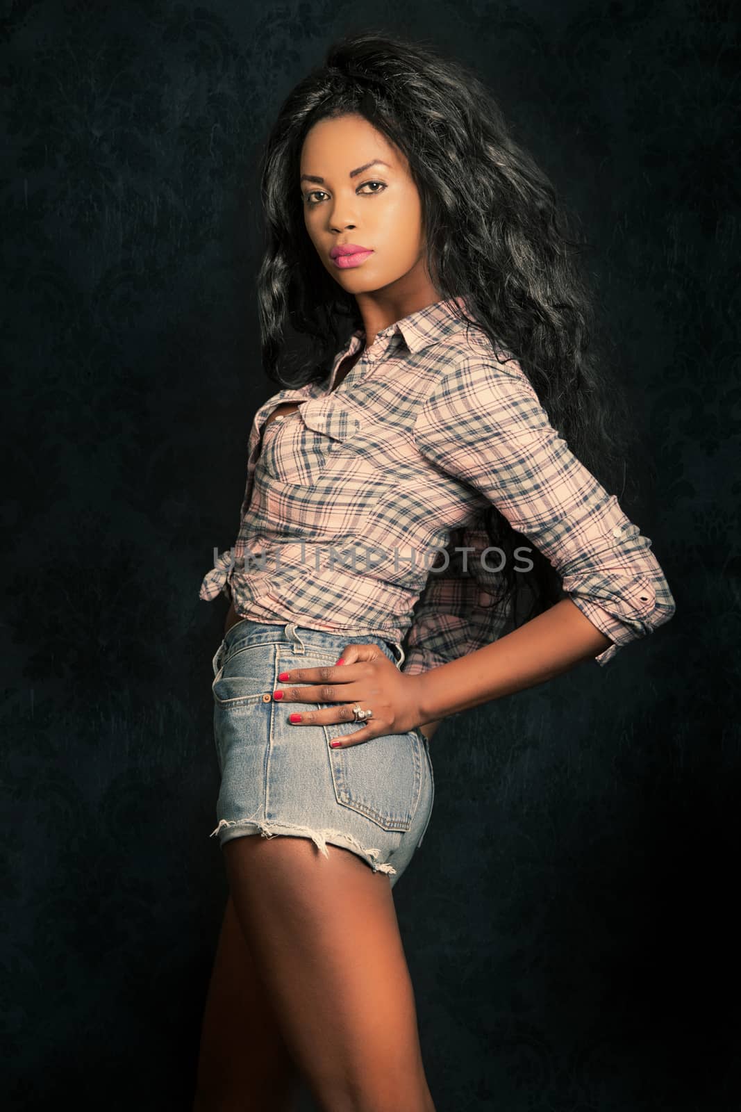 Beautiful young black woman. Afro model with dark background. Casual dressed with short jeans and shirt. Blacks long hair. Posing in the studio, sensual and model beauty.