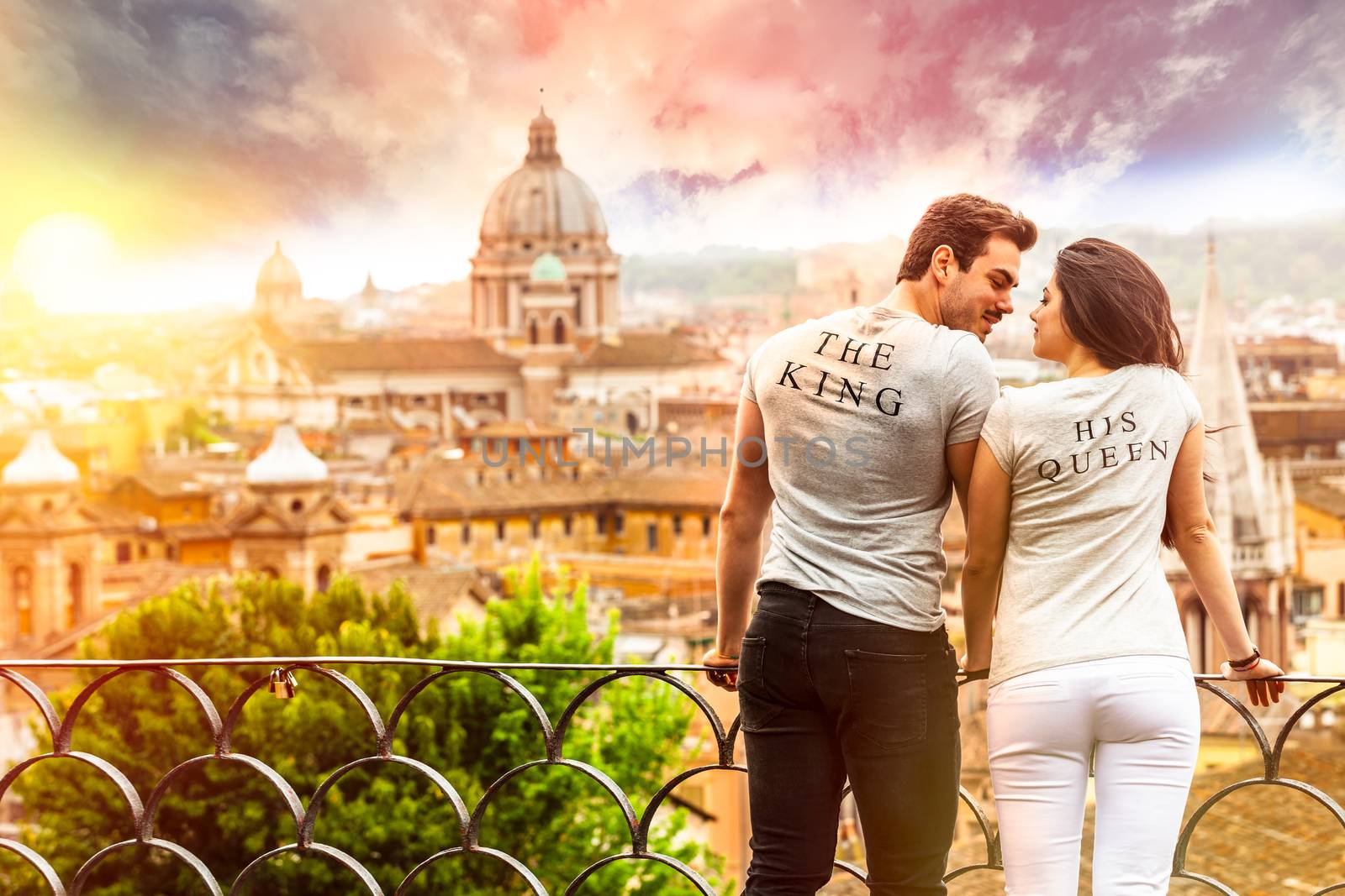 Romantic couple in Rome, Italy. A men and a women, they are looking at each other. On a terrace with railing overlooking Rome at the sunrise. Wearing a gray T-shirt with the words The King and His Queen.