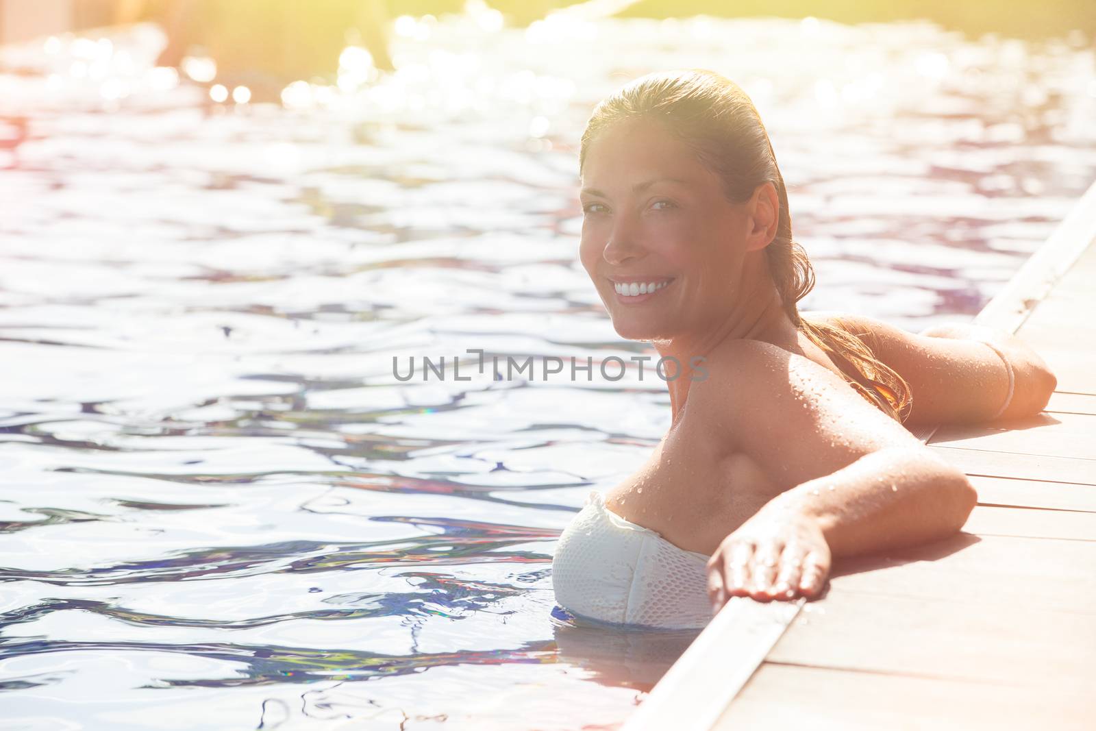 Woman relaxing in the pool water. A beautiful woman with big smile in water in a swimming pool. Relaxation and peace. Against the side of the pool. Intense summer lights.