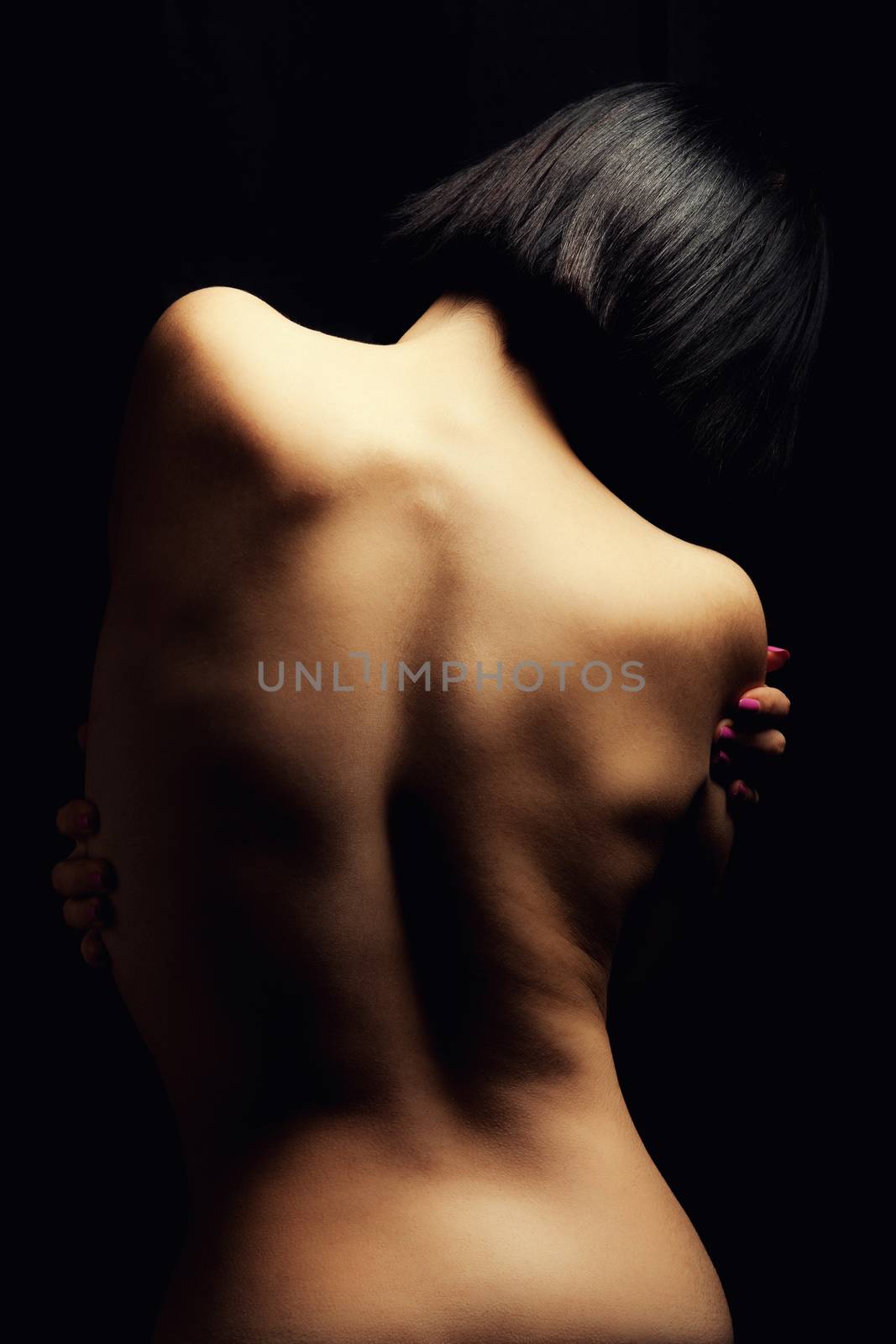 Back of woman. Portrait of the naked back of a young woman. Bobbed hair. Light striking. Smooth skin. Black background.