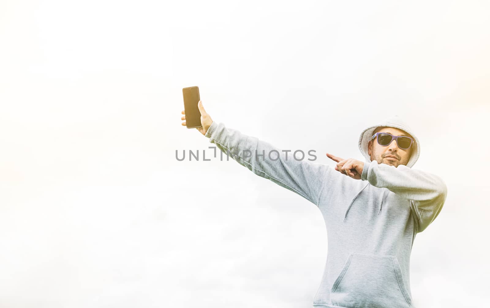 Young man in a grey sweatshirt with blue sunglasses on a white background and pointing at his mobile phone