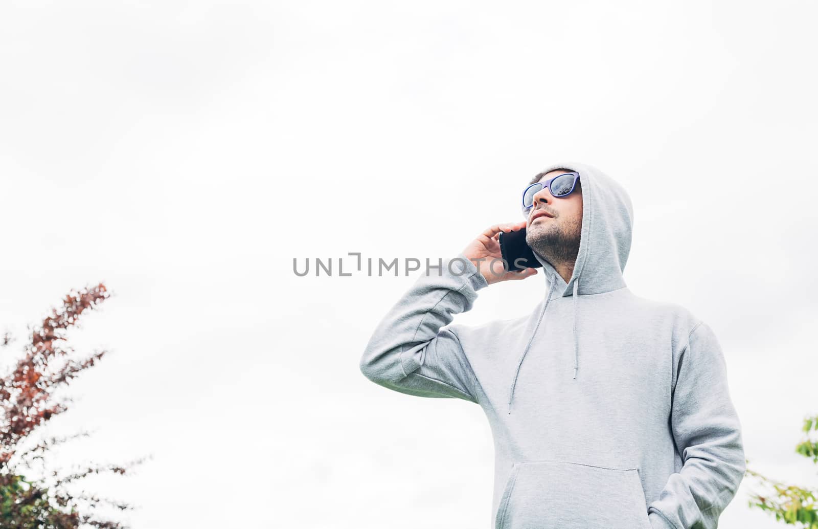 Young man in gray sweatshirt and blue sunglasses holding cell phone in his hands and smiling on a white background