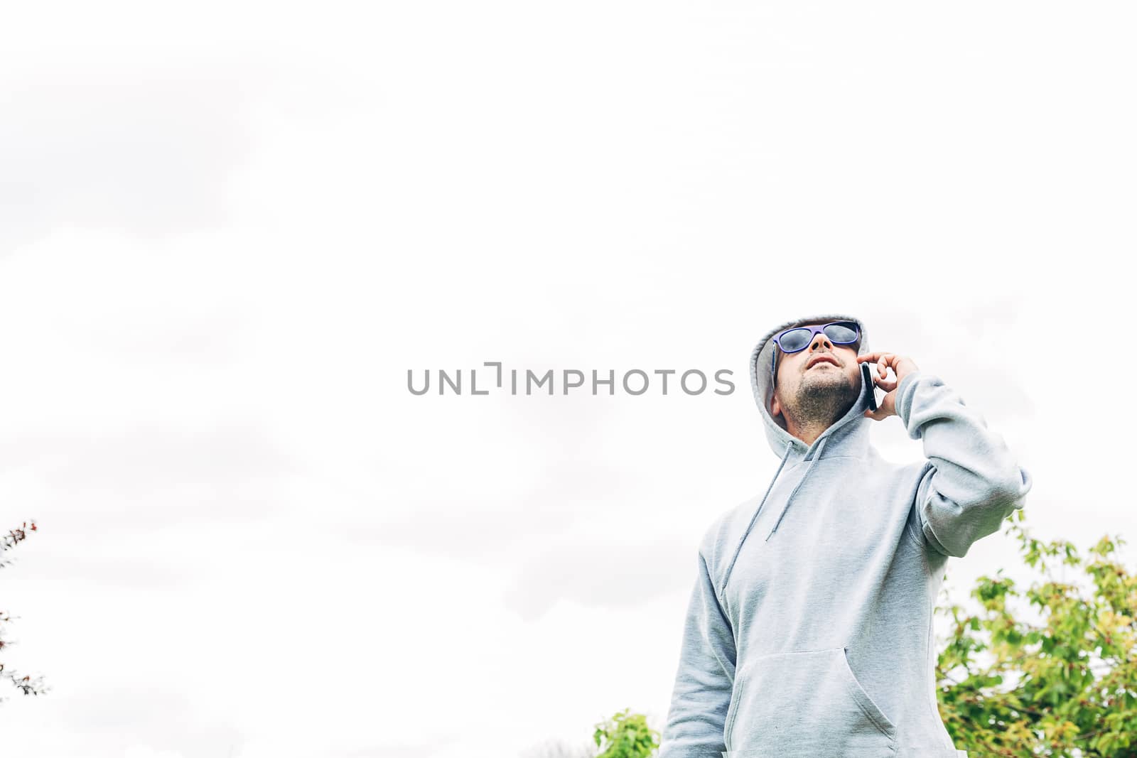 Young man in gray sweatshirt and blue sunglasses holding cell phone in his hands looking up