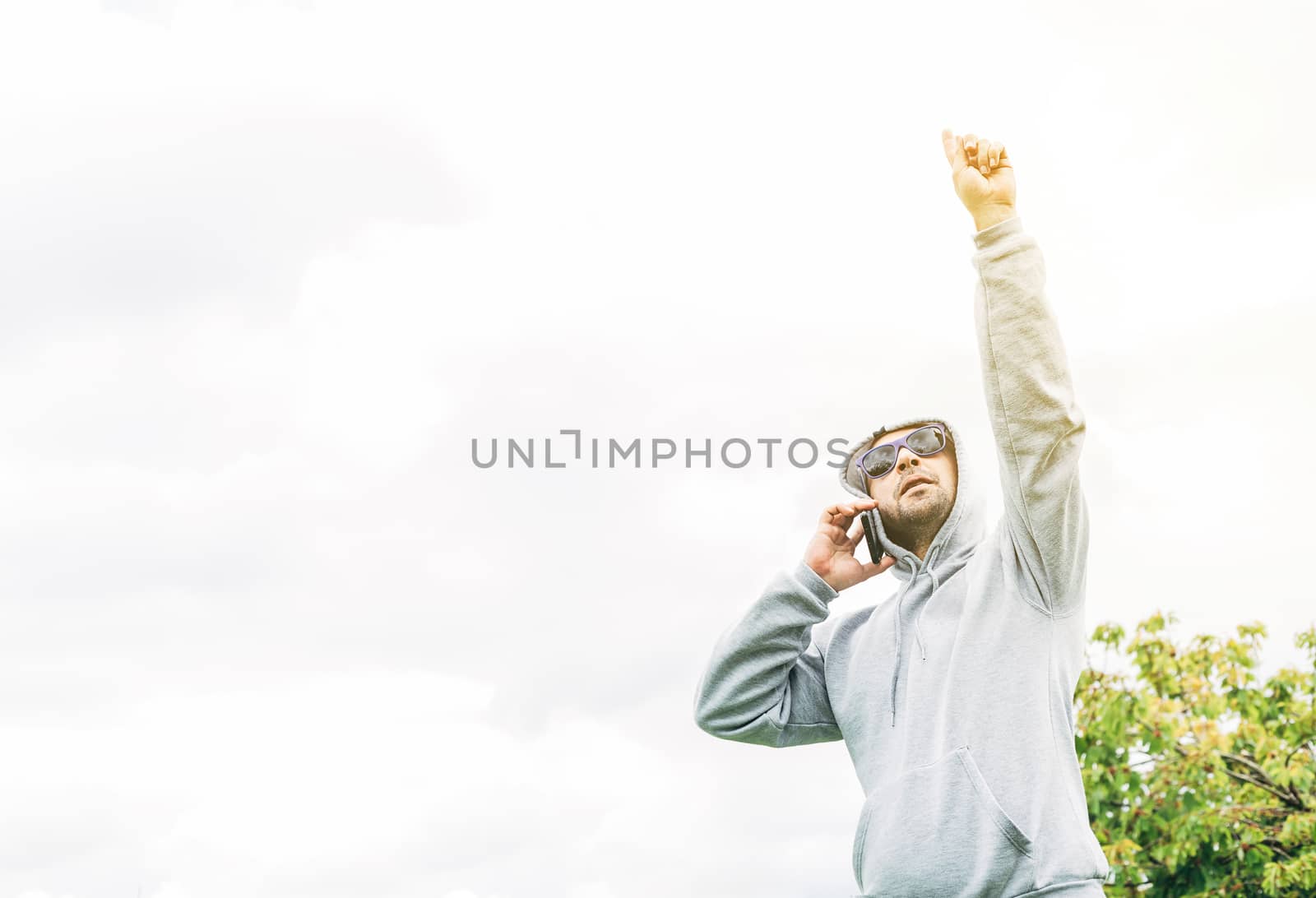 A young A young man with blue sunglasses, gray sweatshirt and beard talking on a cell phone looking up