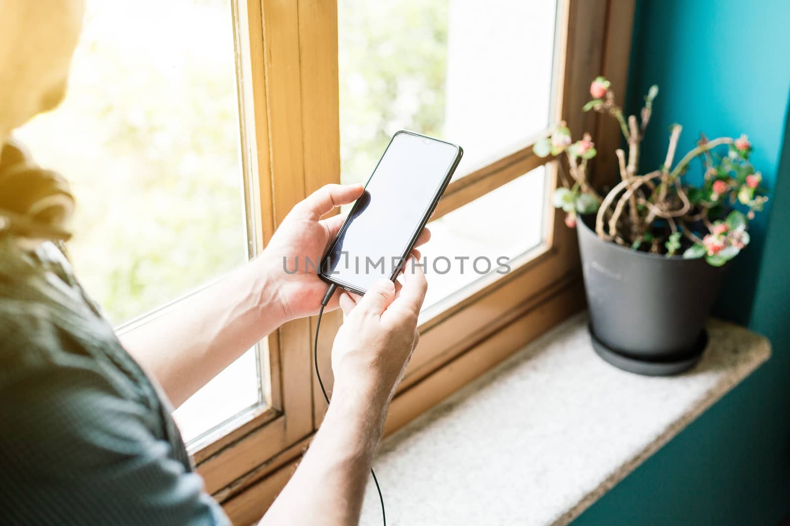 Man holding mobile in his hands next to a window with headphones attached and the sun is shining