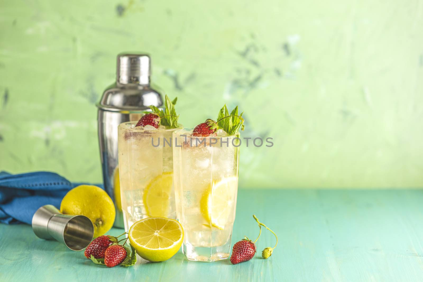 Two glasses of cold icy refreshing drink with lemon and strawberry served with bar tools on green wooden table. Fresh cocktail drinks with ice fruit and herb decoration.
