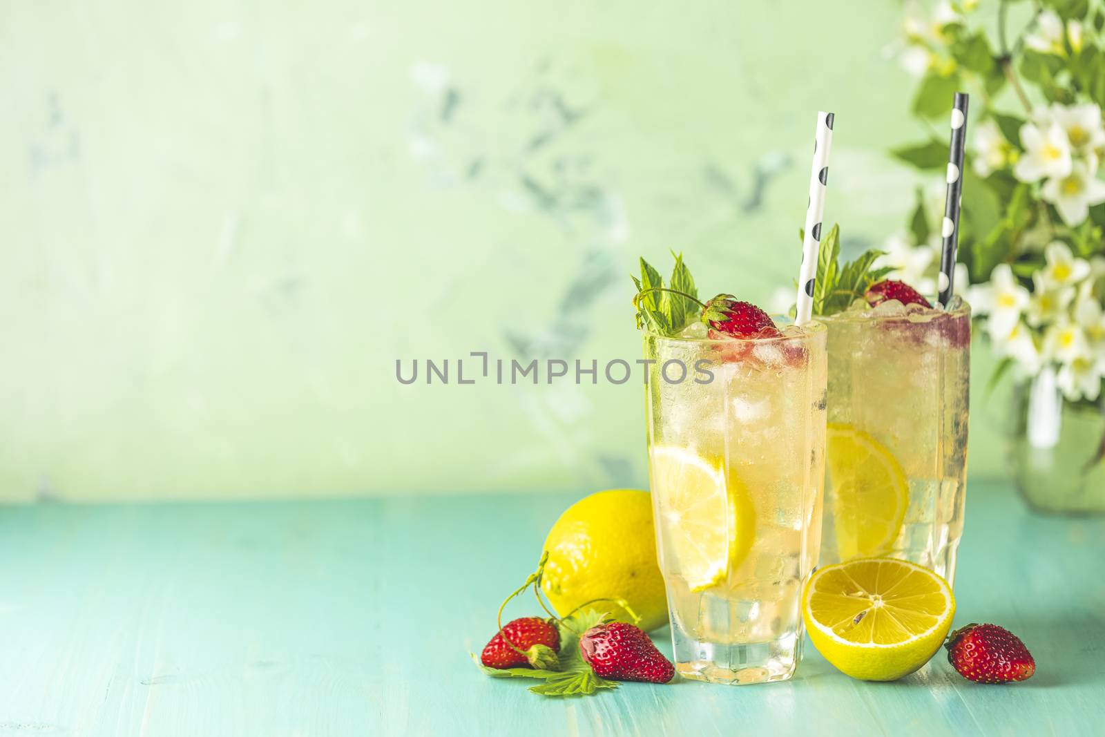 Two glasses of cold icy refreshing drink with lemon and strawberry served with bar tools on green wooden table with white blooming flowers. Fresh cocktail drinks with ice fruit and herb decoration.