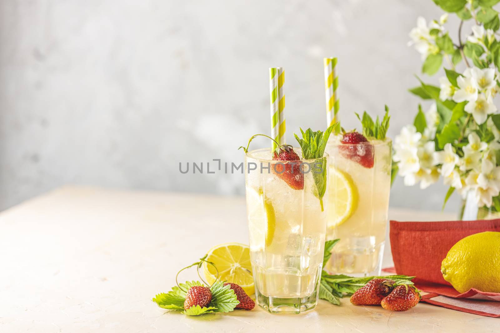 Two glasses of cold icy refreshing drink with lemon and strawberry served with bar tools on light pink table with white blooming flowers. Fresh cocktail drinks with ice fruit and herb decoration.