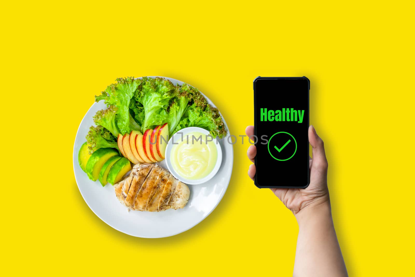 Healthy food concept. Hand holding smartphone with green text healthy with chicken salad and fresh lettuce, avocado, and apple sliced on white plate served with salad dressing on yellow background. 