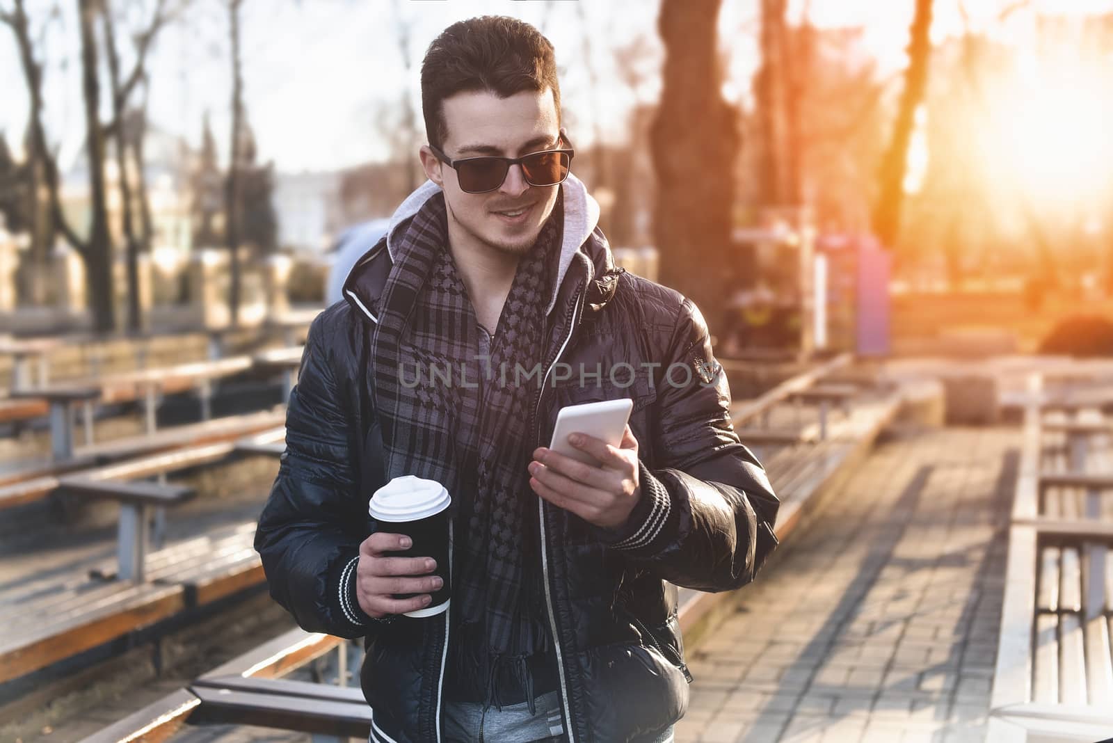 Portrait of attractive man in wearing jacket using mobile phone and holding takeaway coffee while walking through city street.