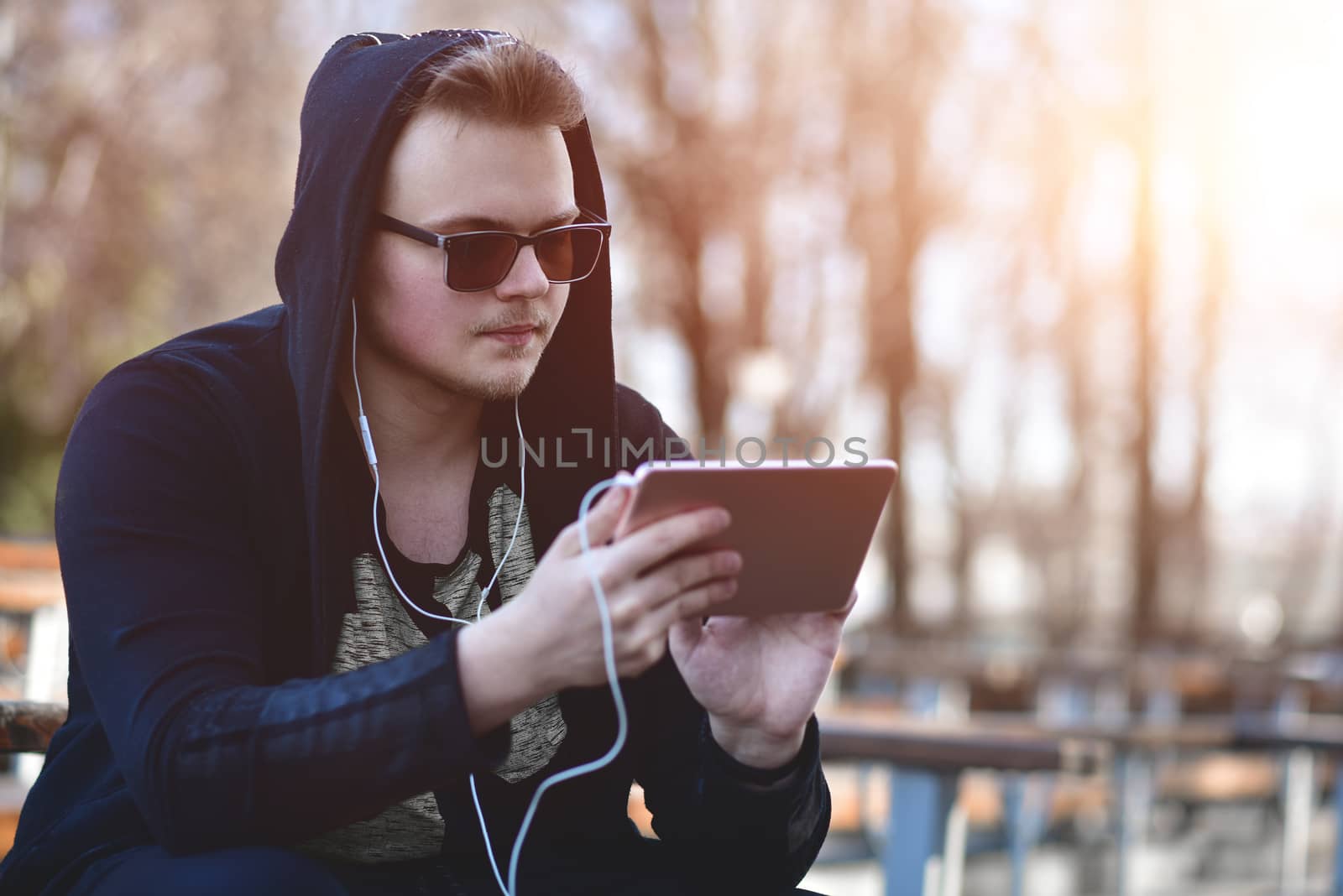 The Man in white headphones and black hood works and communicates on a tablet computer in the park. Closeup of a guy in a black hood and a tablet in his hands running on a tablet.