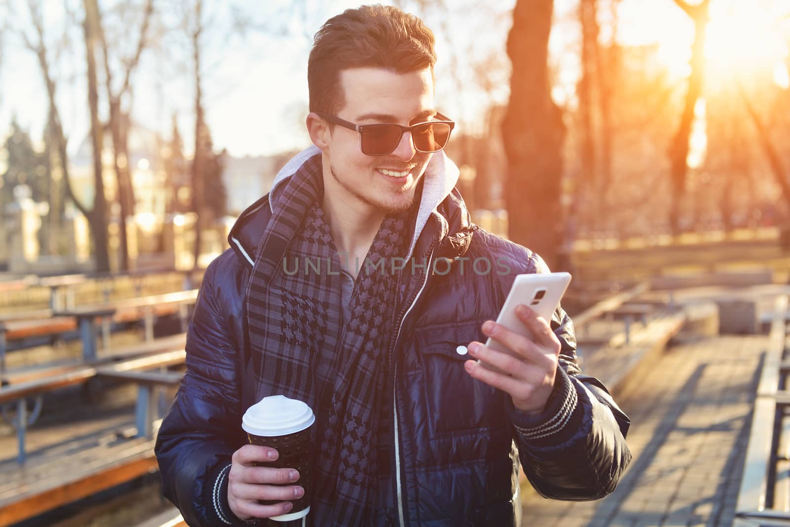 Portrait of attractive man in wearing jacket using mobile phone and holding takeaway coffee while walking through city street.