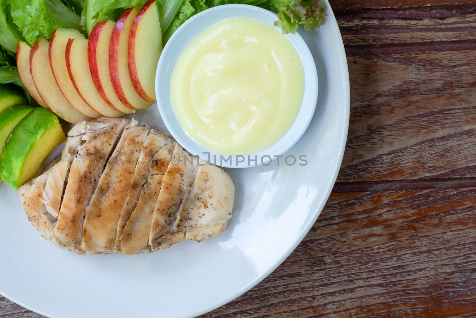 Top view grilled chicken breast steak with sliced avocado, red apple and green lettuce leaves served with salad dressing in white bowl on dish in cafe and restaurant.