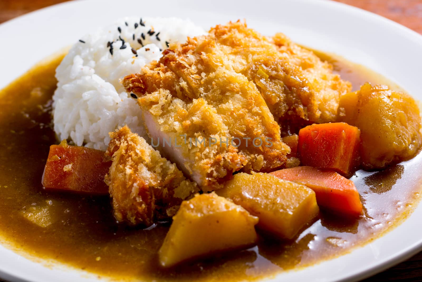 Japanese curry with rice, black sesame, carrot, potato and deep fries chicken in white dish on wooden table.