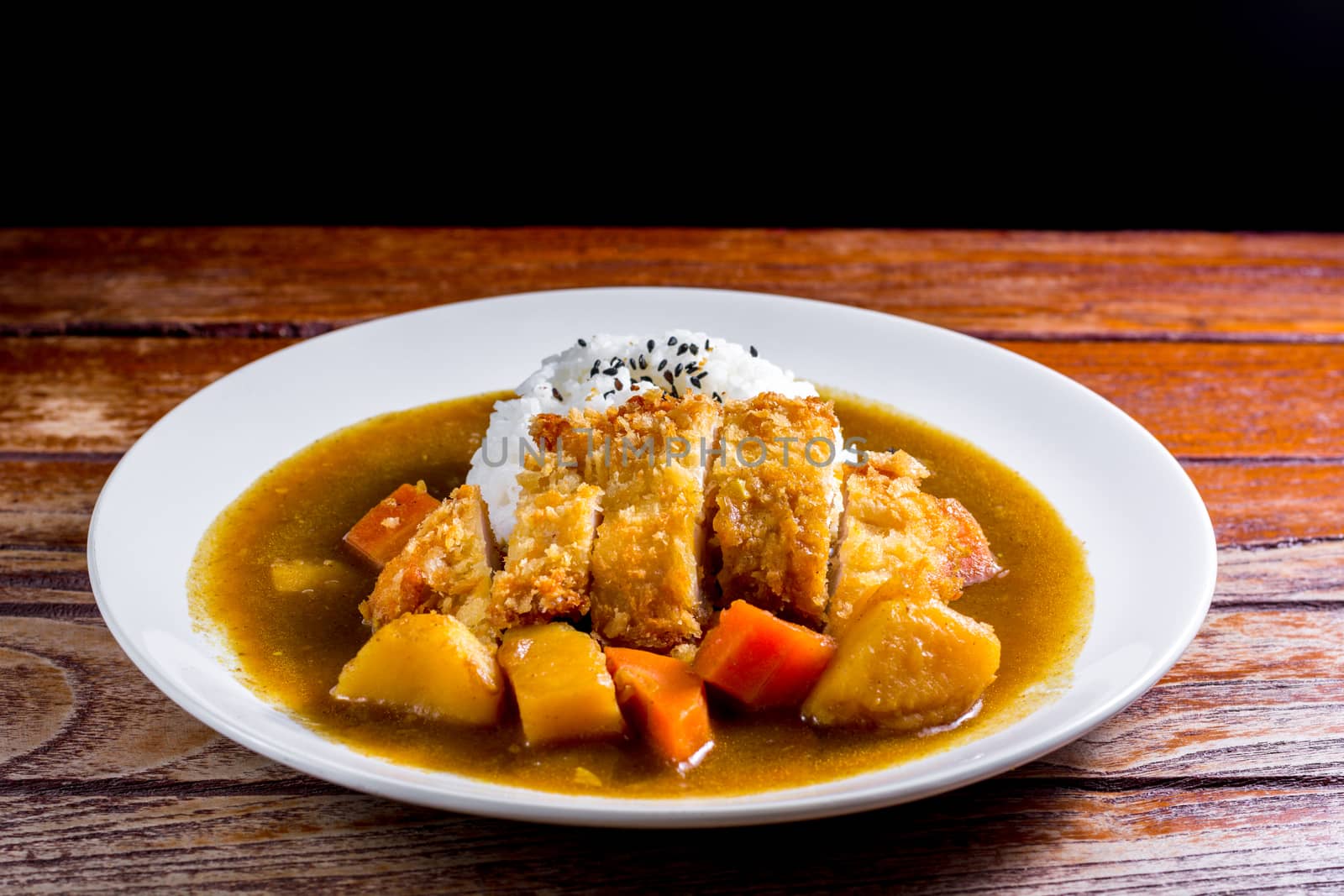 Japanese curry with rice, black sesame, carrot, potato and deep fries chicken in white dish on wooden table.