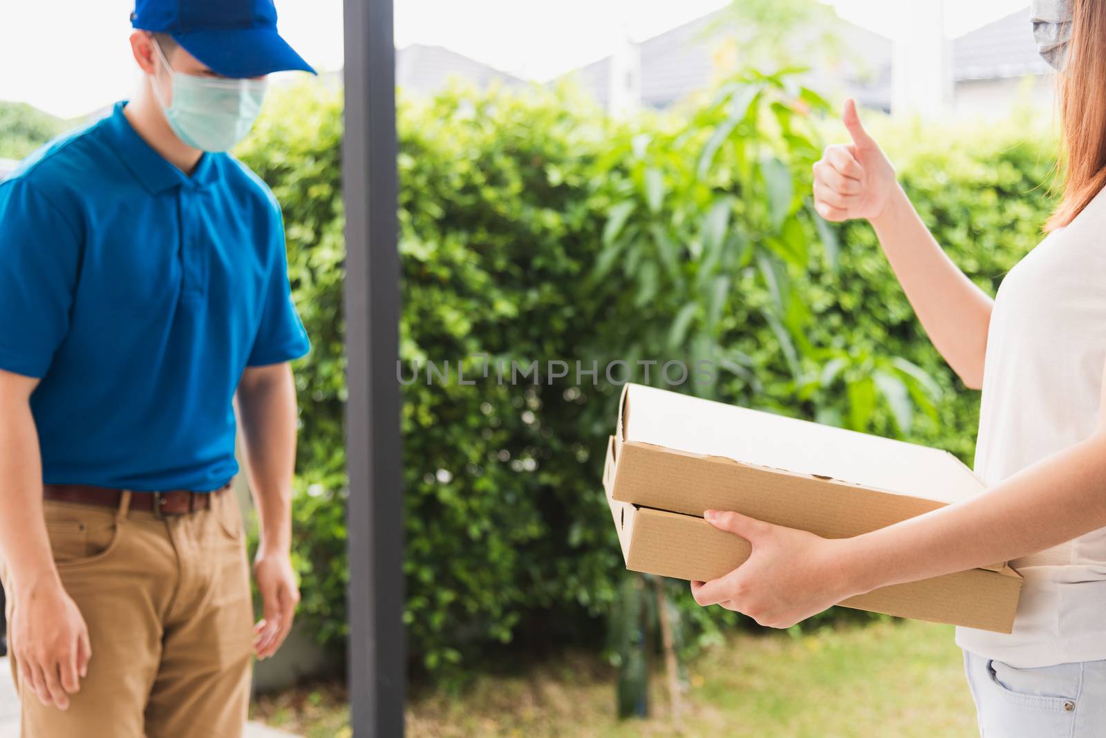 Asian delivery courier young man giving food pizza box to woman customer receive both protective face mask and show thumb up finger for good job, under curfew quarantine pandemic coronavirus COVID-19