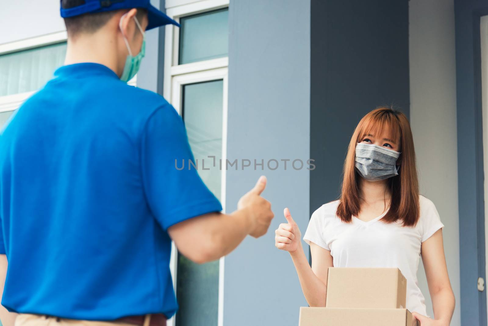 Asian delivery express courier young man giving parcel boxes to woman customer receiving both protective face mask and show thumbs up finger for good support sign, under curfew pandemic coronavirus