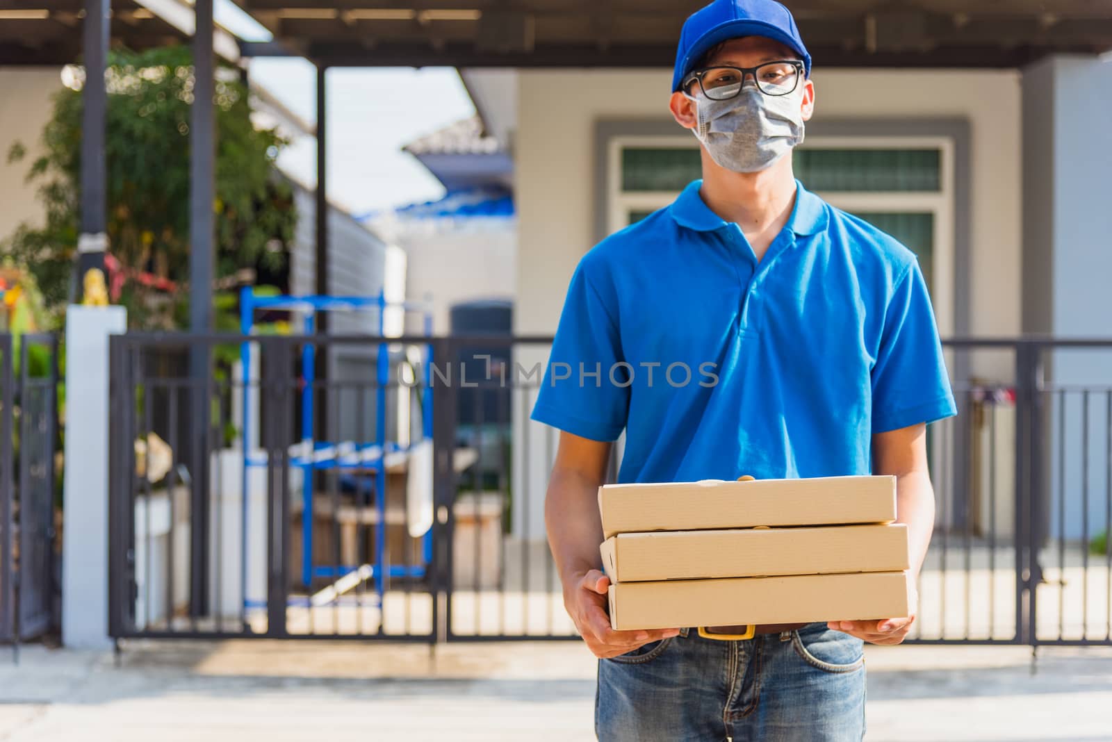 Asian Young delivery man courier online with food order is pizza boxes in uniform he protective face mask, service customer in front of the house under curfew quarantine pandemic coronavirus COVID-19