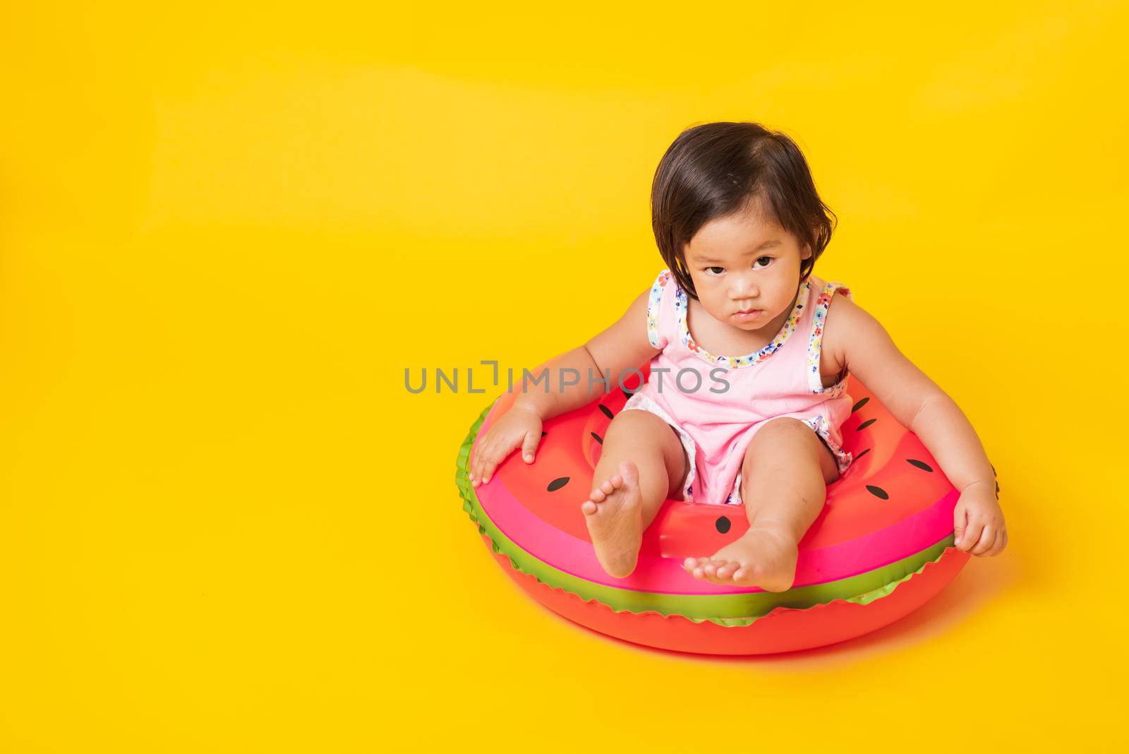 Summer vacation concept, Portrait Asian happy cute little baby girl wear swimsuit sitting in watermelon inflatable ring, Kid have fun sit in inflatable, studio shot isolated yellow background