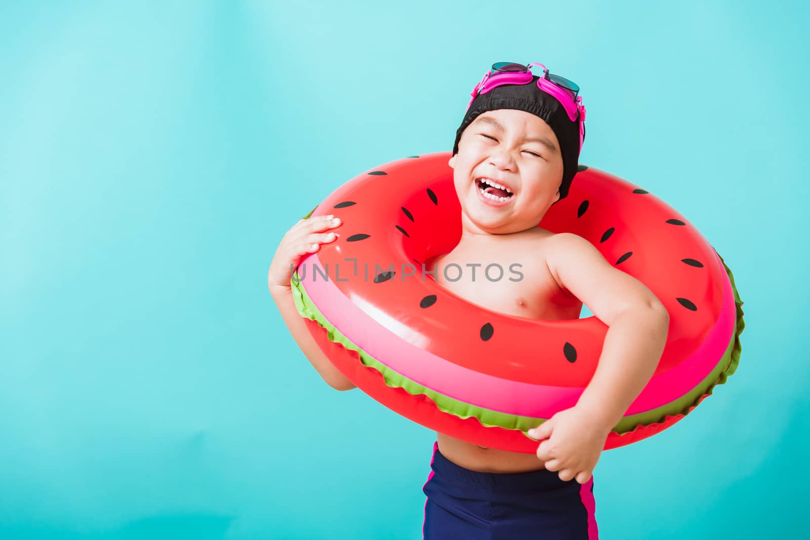 Summer vacation concept, Portrait Asian happy cute little child boy wear goggles and swimsuit hold watermelon inflatable ring, Kid having fun on summer vacation, studio shot isolated blue background