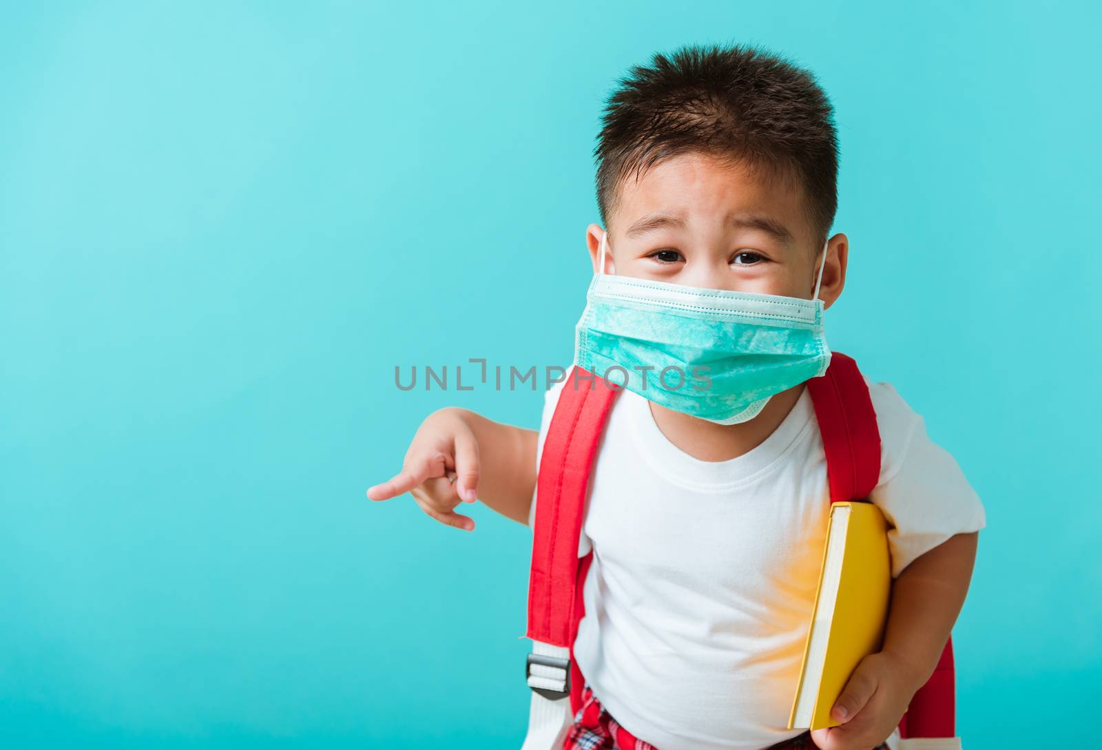 Back to school coronavirus Covid-19 education. Portrait Asian little child boy kindergarten wear face mask protective and school bag hold book before going to school pointing to side away, studio shot