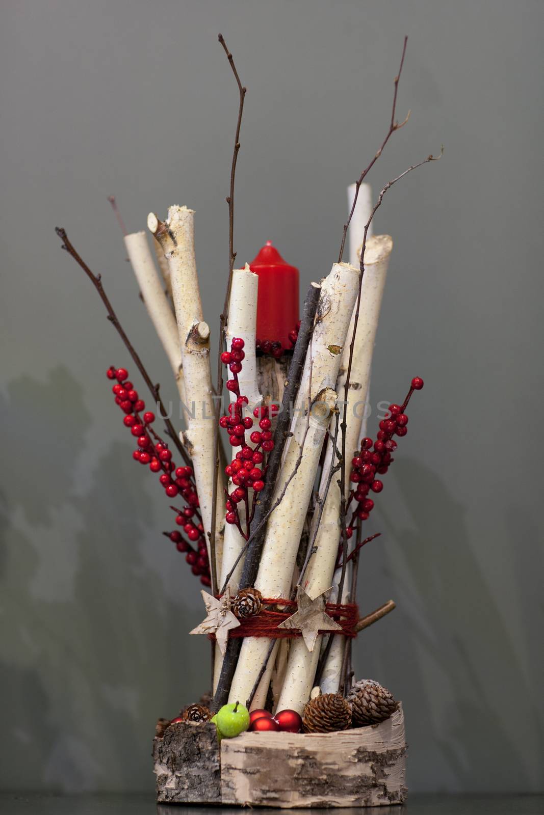 Christmas red candle decorated with a bunch of wooden twigs and berries and fir cones on a gray background