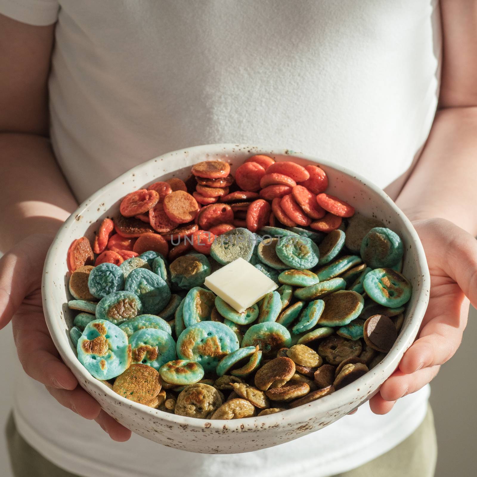 Trendy food - pancake cereal. Colorful mini cereal pancakes in bowl in kids hands. Child hold tiny pancakes with natural colorant - green matcha, turquose spirulina, blue pea, red dragon fruit