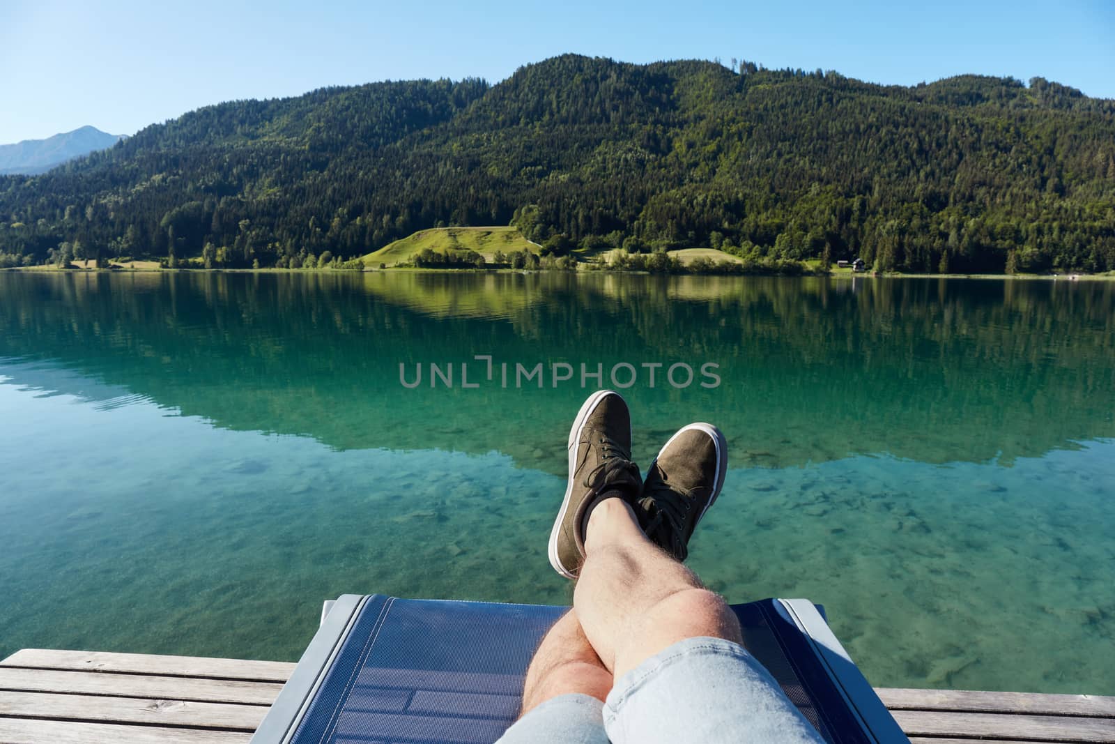 The legs of a man sitting on a deckchair, on the shore of a lake