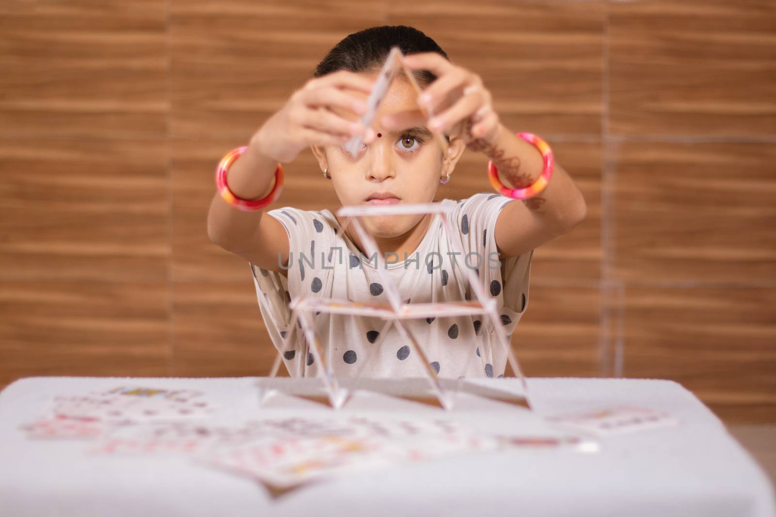 Young girl kid busy in building Pyramid with house of cards at home - concept of focus or concentration excerise game for school children by lakshmiprasad.maski@gmai.com