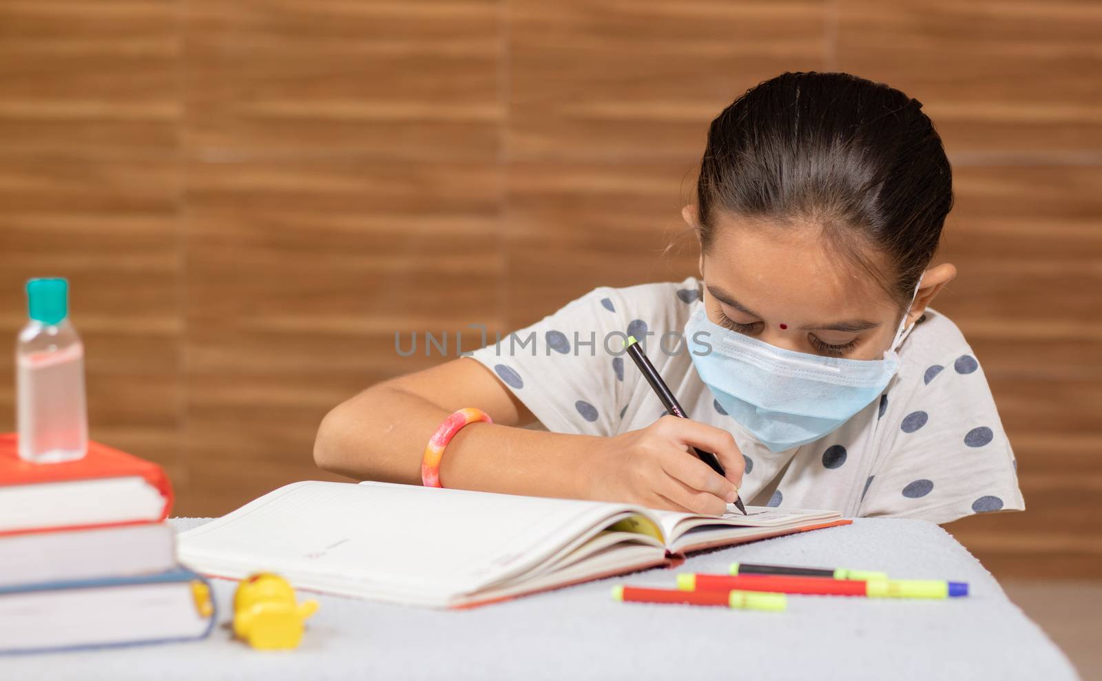 Concept of homeschooling, young girl with medical mask busy in writing at home during covid-19 or coronavirus pandemic lock down. by lakshmiprasad.maski@gmai.com