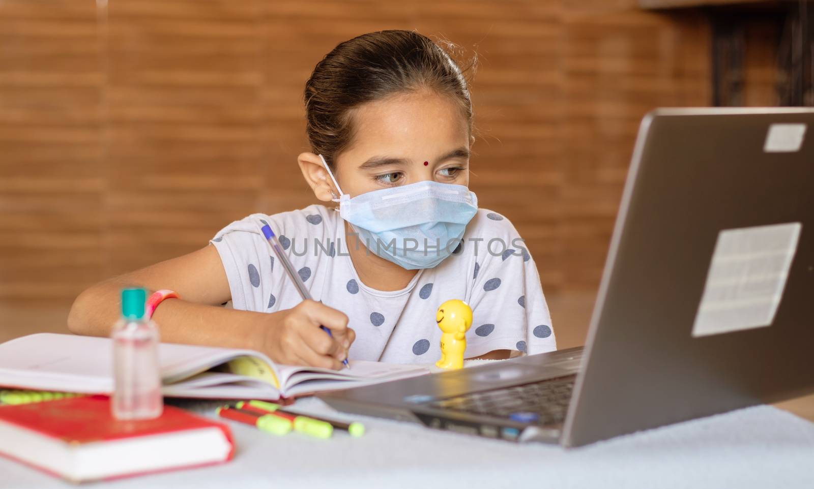 Concept of homeschooling and e-learning, young girl with medical mask writing by looking into laptop during covid-19 or coronavirus pandemic lock down