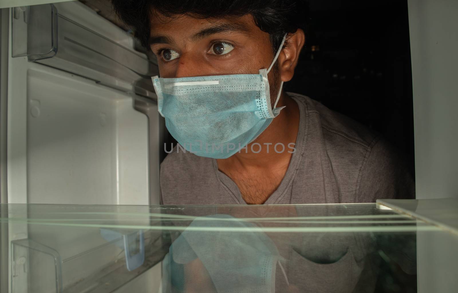 Man in a medical mask looks into empty fridge or refrigerator for food - Concept of no pantry food available during home quarantine at covid-19 or coronavirus