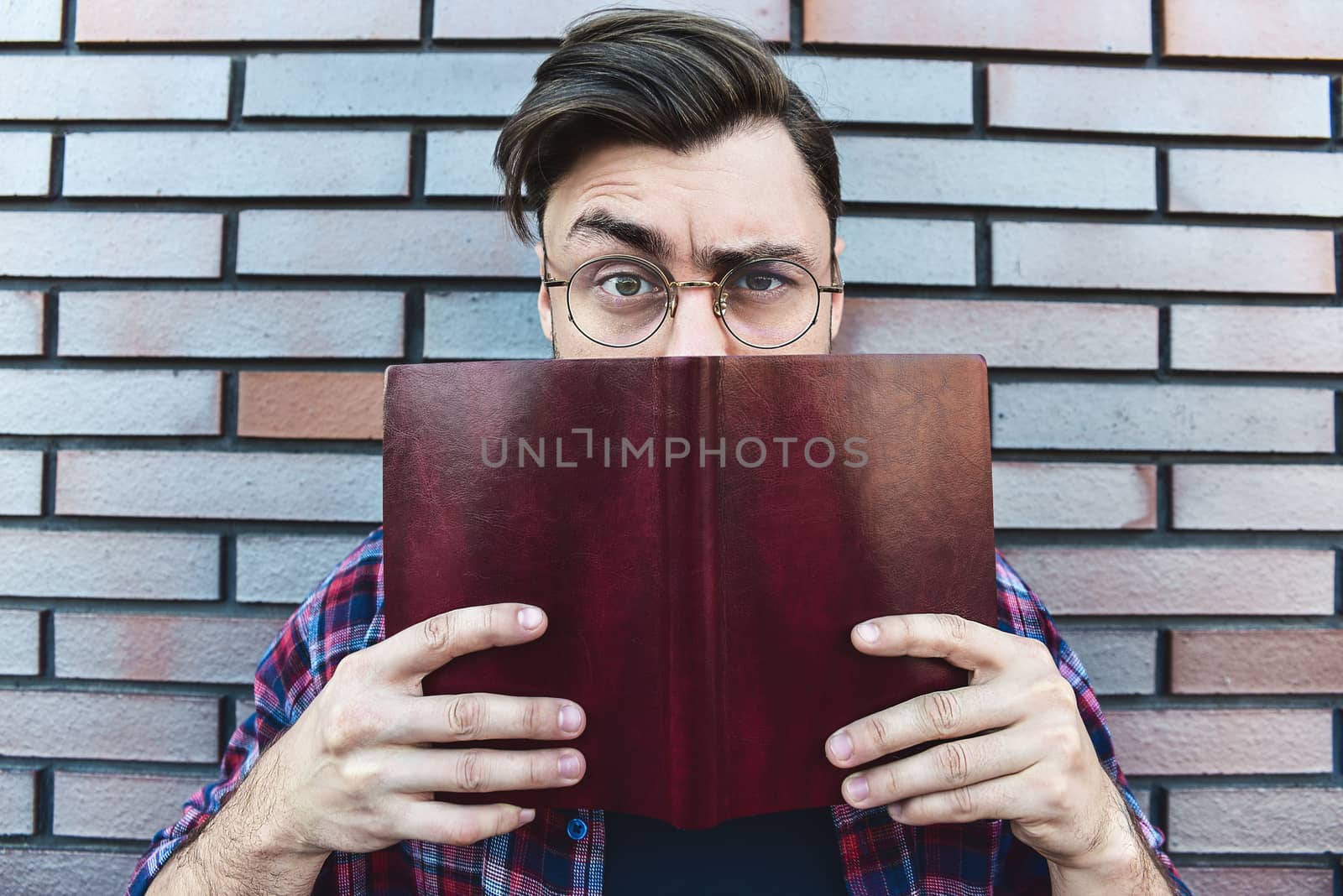 Funny face. Young hipster guy wearing eyeglasses with picked up one browe looking at camera and holding a book or note book on brick wall background.