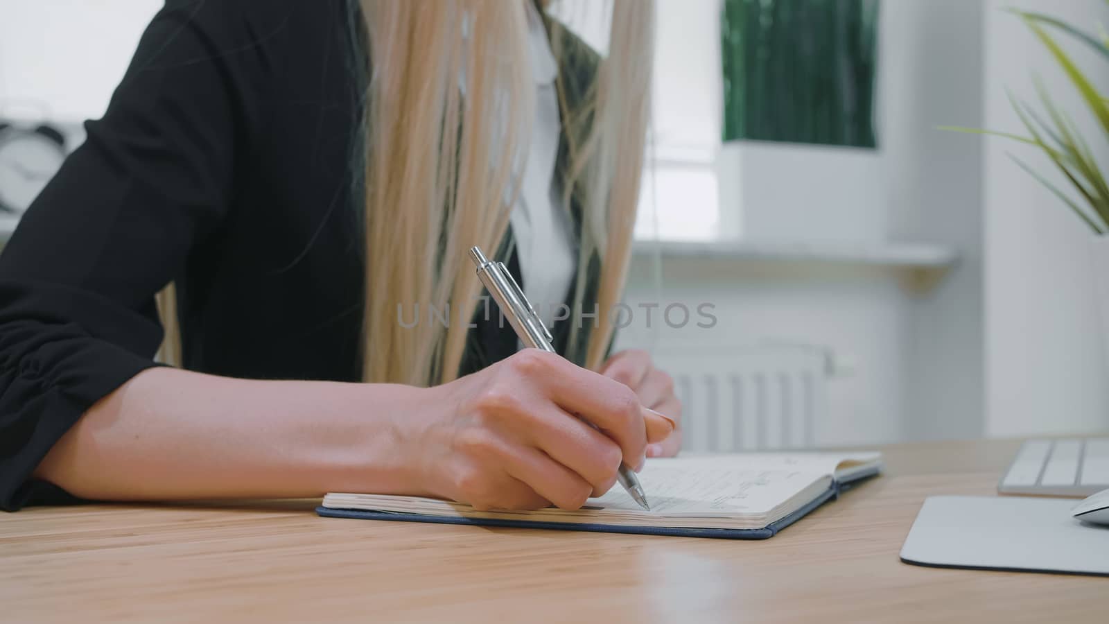 Crop view of woman with long blond hair in bright casual shirt, sitting at wooden desk and writing down information with shiny metal pen into daily planner at day.